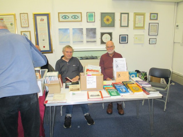 Nigel Wassell and Paul Reynolds sit behind the SWWIAS table with a selection of books, at the Local History Book Fair